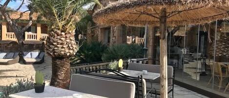 An outdoor seating area with tables and chairs, surrounded by palm trees and a thatched umbrella.