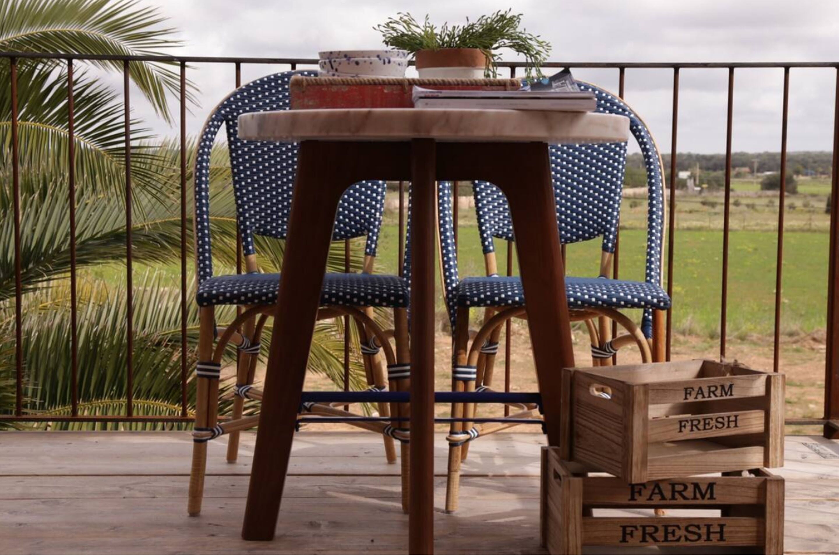Una mesa de madera con sillas y cajas etiquetadas como "FARM FRESH" en un balcón con vista a un campo verde.
