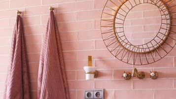 Un baño con pared de ladrillo rosa, un espejo redondo, un aparador antiguo con diseños florales y dos toallas rosas colgadas de ganchos.