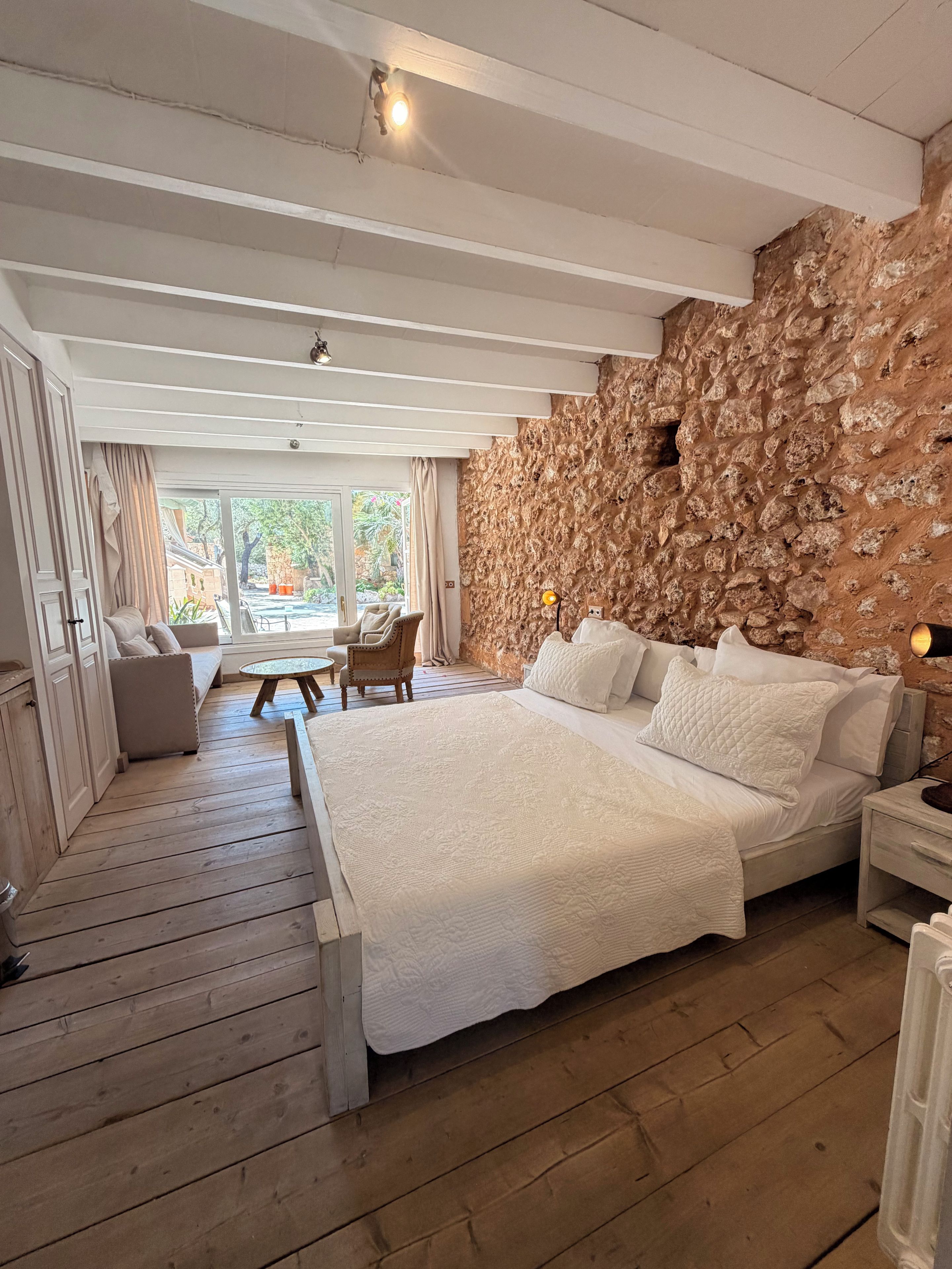 Une chambre avec un grand lit, un plancher en bois, un mur en pierre et une vue sur l’extérieur.