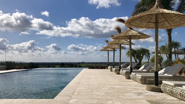 A poolside area with lounge chairs and thatched umbrellas, a clear sky, and a distant view of a landscape. - Hotel Rural Es Turó (Ses Salines)