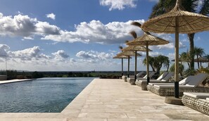 A poolside area with lounge chairs and thatched umbrellas, a clear sky, and a distant view of a landscape.