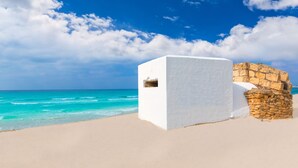 A white building with a stone wall adjacent to a beach with clear turquoise waters and a blue sky with clouds. - Hotel Rural Es Turó (Ses Salines)