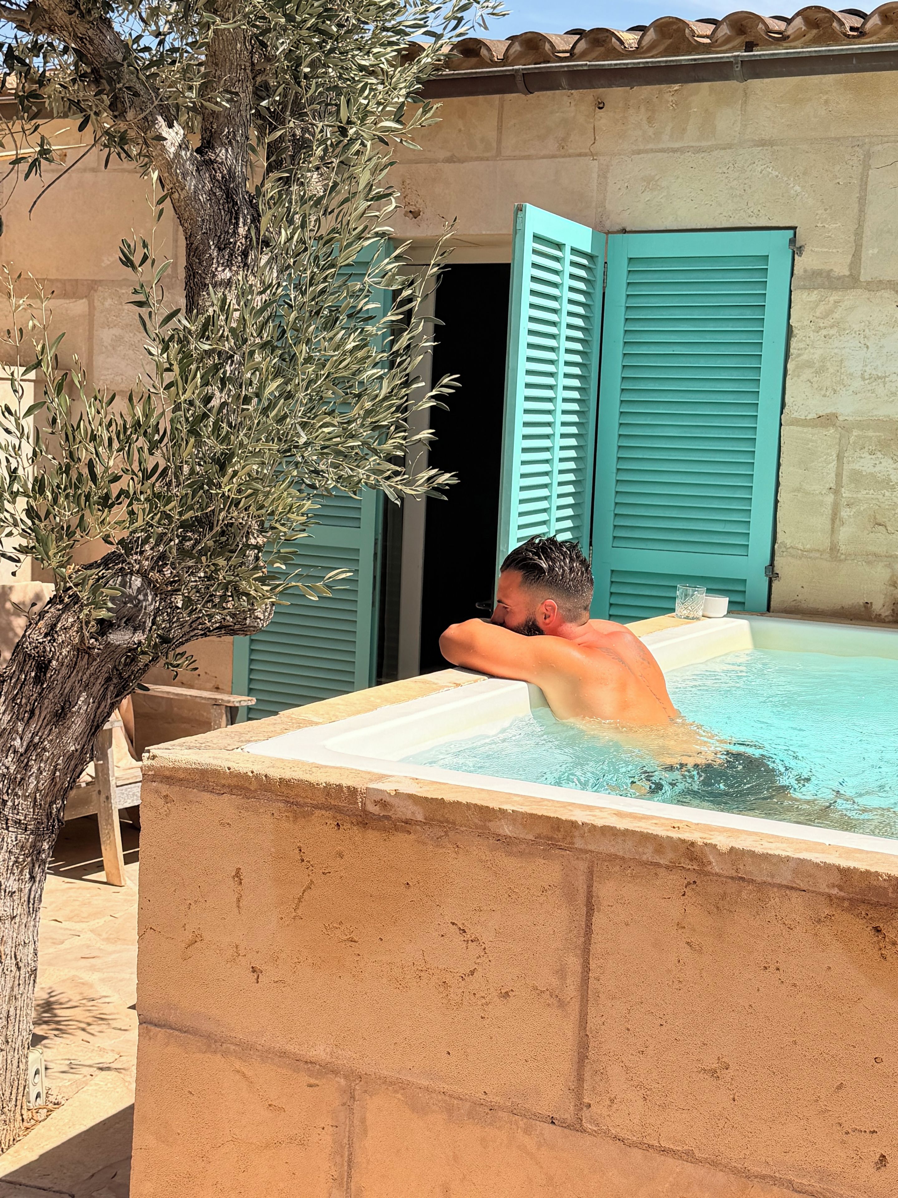 Un hombre se relaja en un jacuzzi, con un árbol y un edificio al fondo.