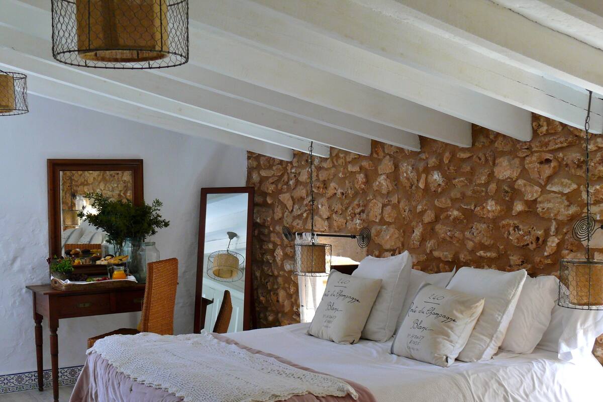 Quarto com parede de pedra, cama com roupa de cama branca, escrivaninha de madeira e um espelho.