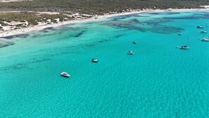 An aerial view of a coastal area with clear turquoise waters, several boats, and a beach with buildings in the background. - Hotel Rural Es Turó (Ses Salines)