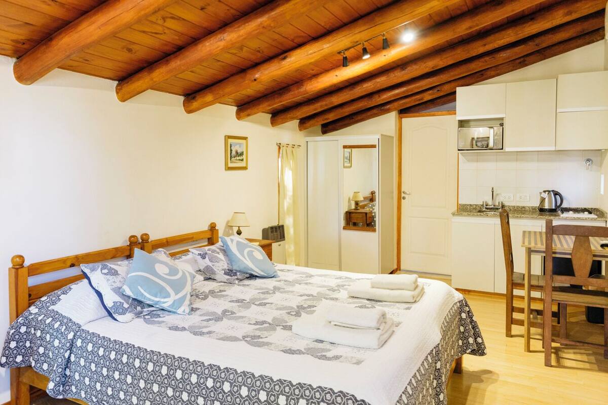 A bedroom with a wooden ceiling, a bed with patterned bedding, a bedside table with a lamp, and a view of a kitchen with a microwave and a kettle.