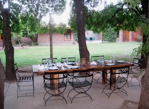 An outdoor dining area with a long table set for six, surrounded by metal chairs, with trees and a brick building in the background.