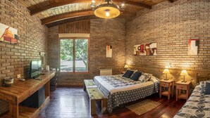A rustic room with exposed brick walls, a wooden ceiling, a large bed with patterned bedding, a desk with a television, and a window with a view of greenery. - Lujan de Cuyo B&B (Luján de Cuyo)