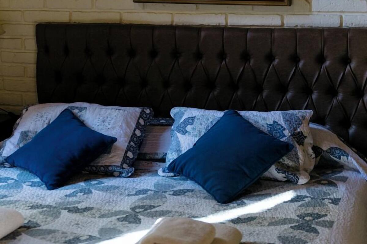 A bedroom with a brick wall, a framed painting of horses, a tufted headboard, and blue pillows.