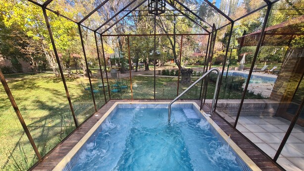 A rectangular hot tub with a metal frame and glass panels, surrounded by a wooden deck and a grassy area with trees and a patio in the background. - Lujan de Cuyo B&B (Luján de Cuyo)