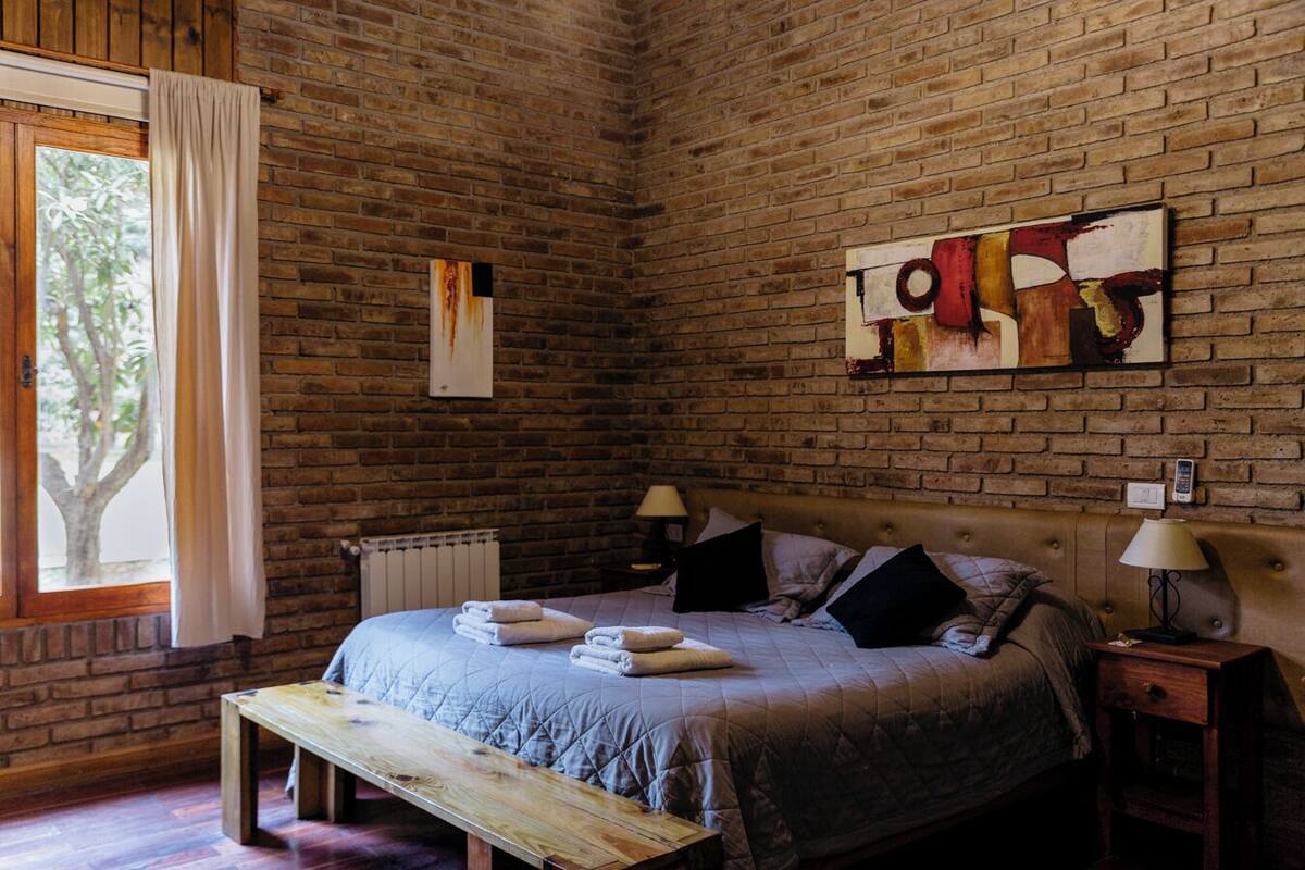 A bedroom with a brick wall, a bed with a quilt, a bench, a radiator, and a window with curtains.