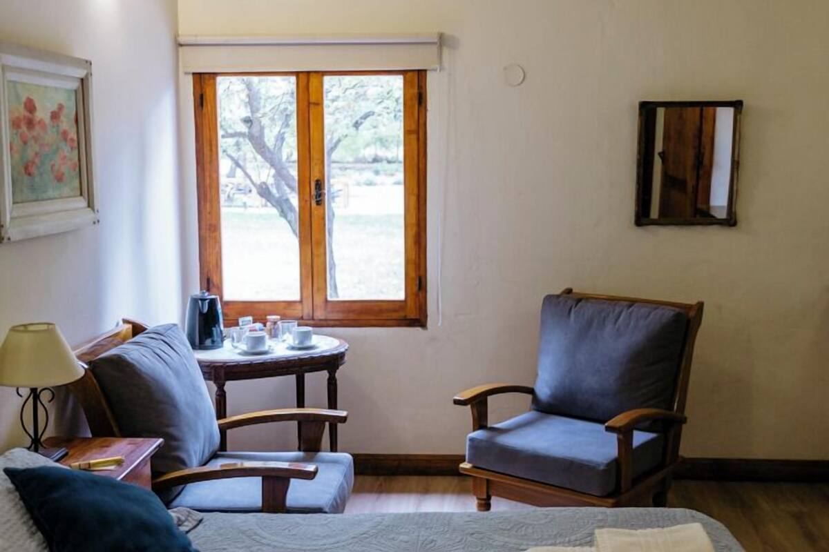 A cozy room with a bed, a chair, a small table with a lamp, and a window with a view of trees.