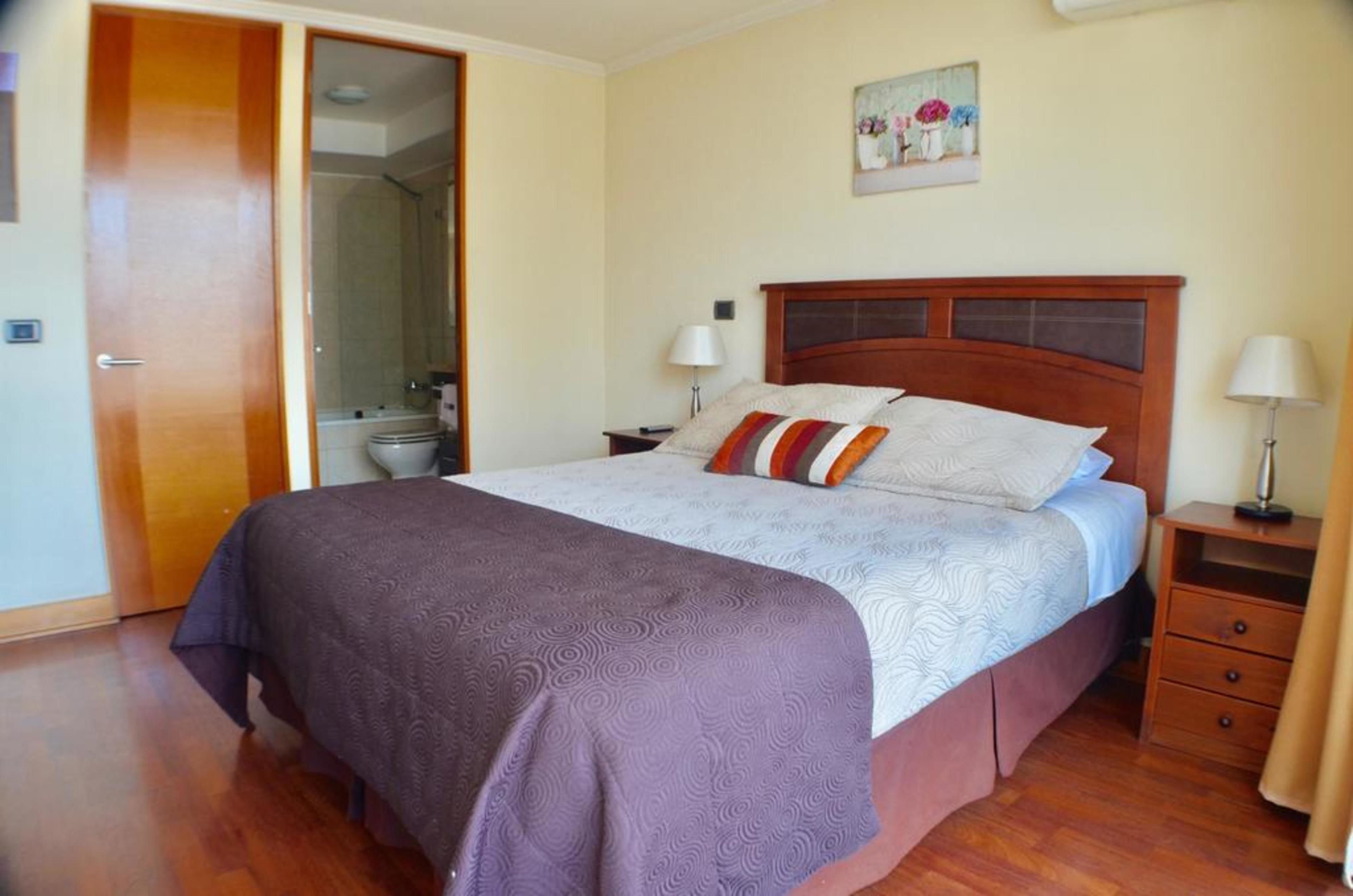 A bedroom with a wooden bed, a nightstand, a bedside table, a door, and a bathroom visible through an open door.