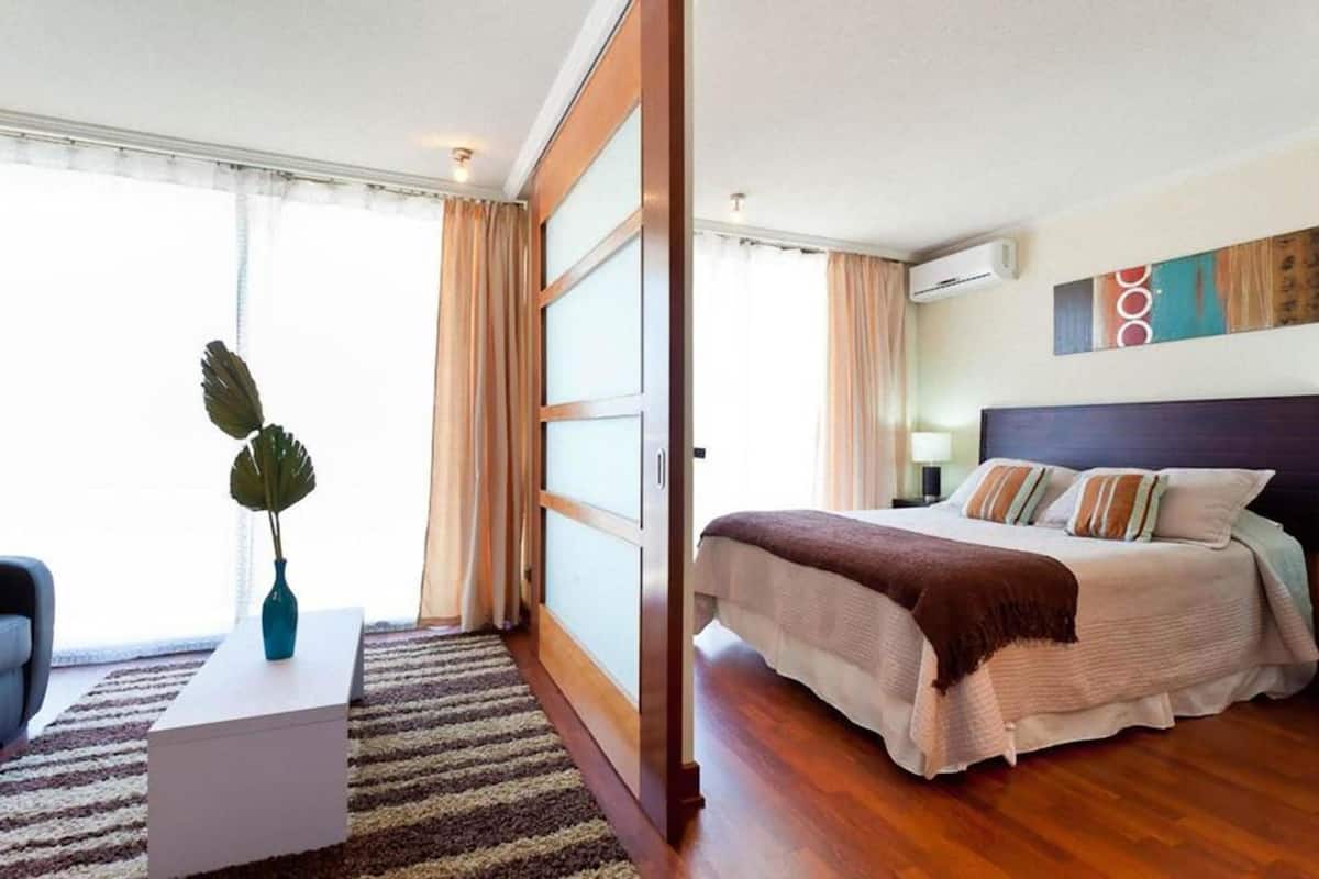 Une chambre d’hôtel avec un lit, une table de chevet blanche ornée d’un vase, une grande fenêtre avec des rideaux et un appareil de climatisation fixé au mur.