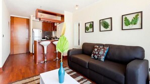 Uma sala de estar com um sofá de couro preto, uma mesa de centro branca e um vaso azul com uma planta. Há quadros com folhas nas paredes.