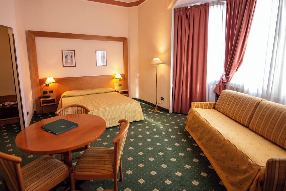 A hotel room with a bed, a sofa, a dining table with chairs, a standing lamp, and a picture on the wall.