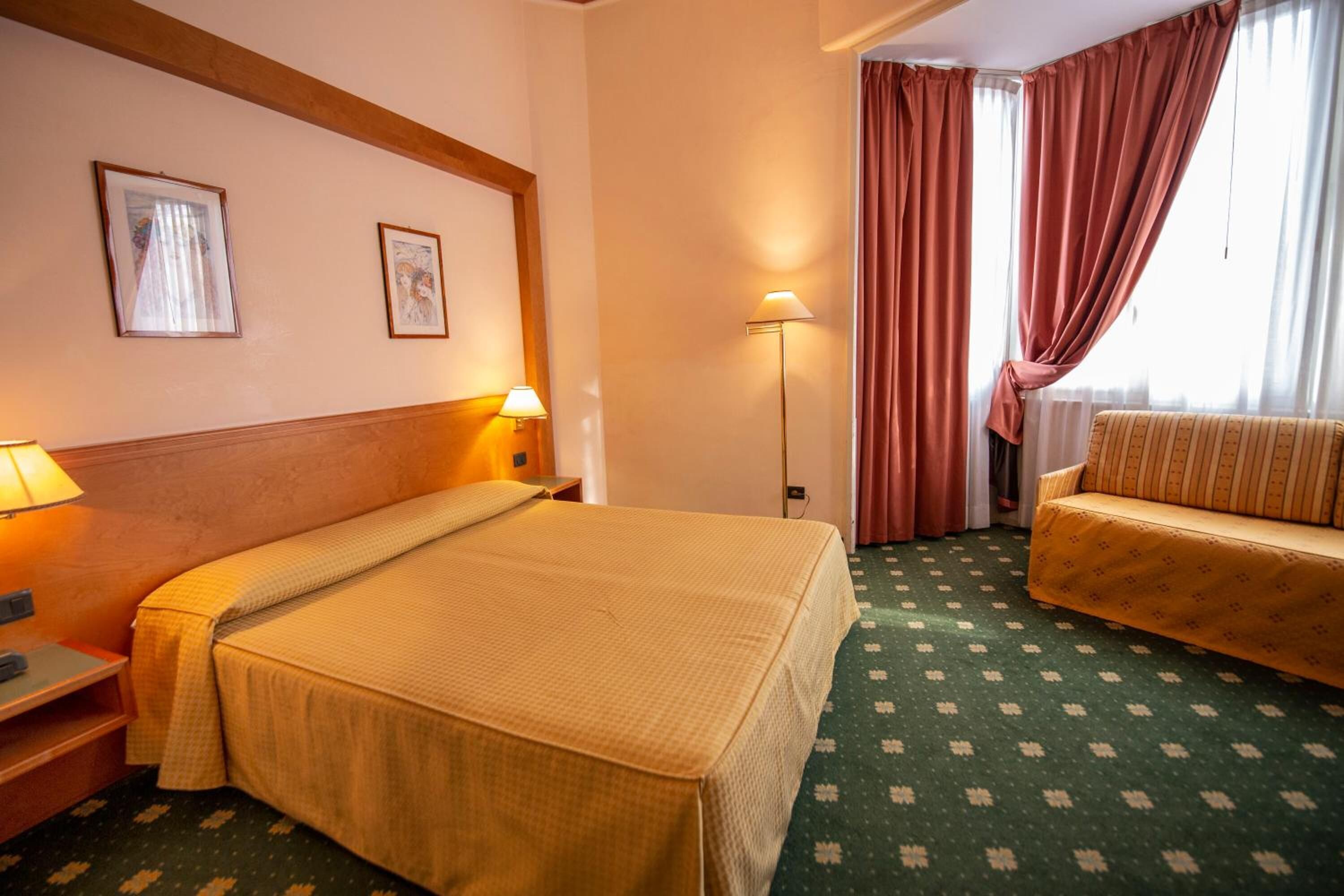 Una habitación de hotel con cama, sofá, dos lámparas de pared y una ventana con cortinas.