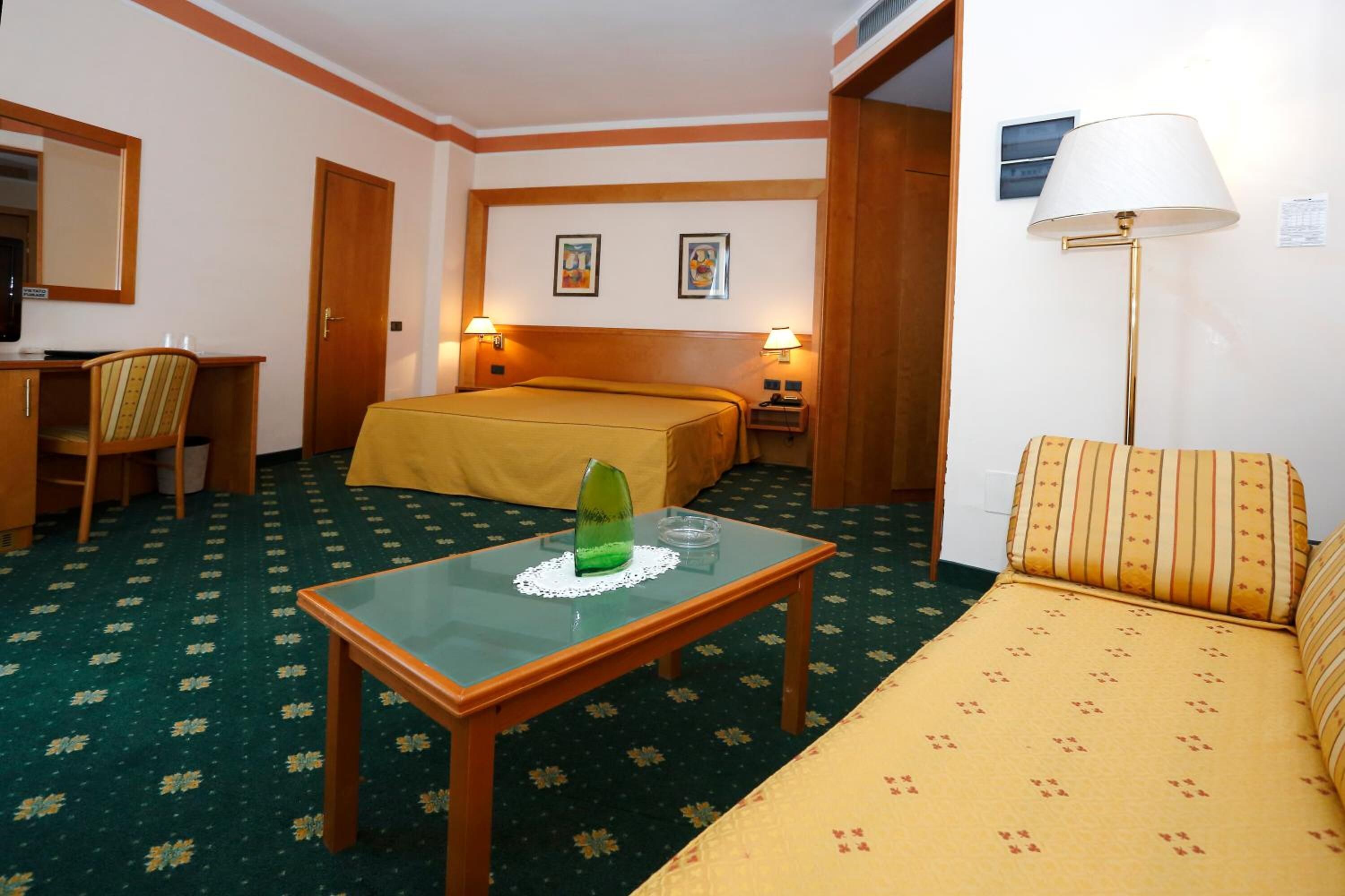 Ein Hotelzimmer mit Bett, Sofa, Schreibtisch mit Stuhl, Tisch mit Glasplatte und zwei Wandleuchten.
