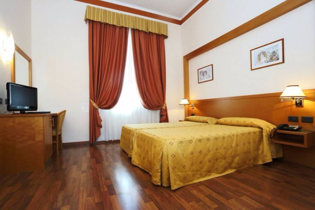 A hotel room with a bed, a desk with a computer, a telephone, and two wall-mounted lamps.