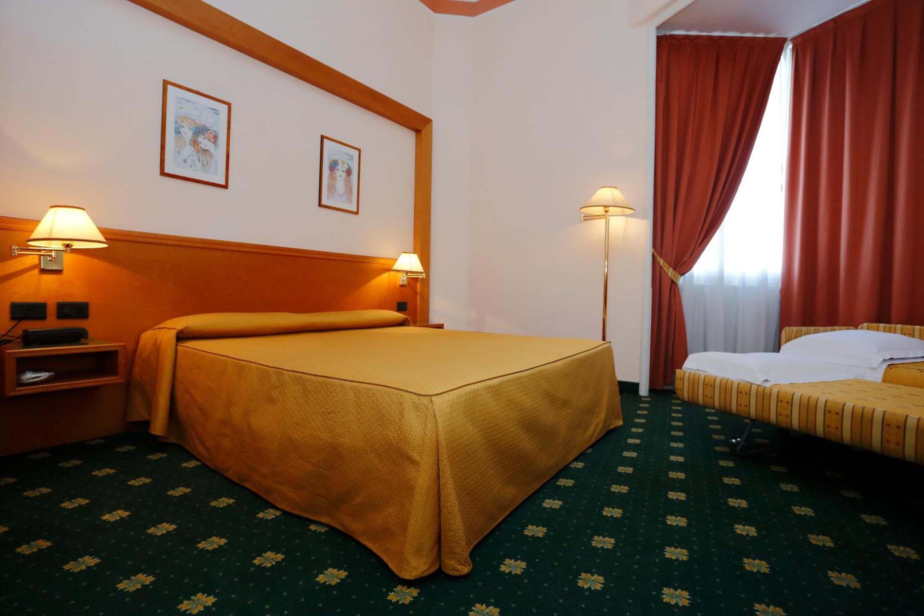 Une chambre d’hôtel avec un lit double, un lit simple, deux tables de chevet, deux lampes fixées au mur et une fenêtre avec des rideaux rouges.