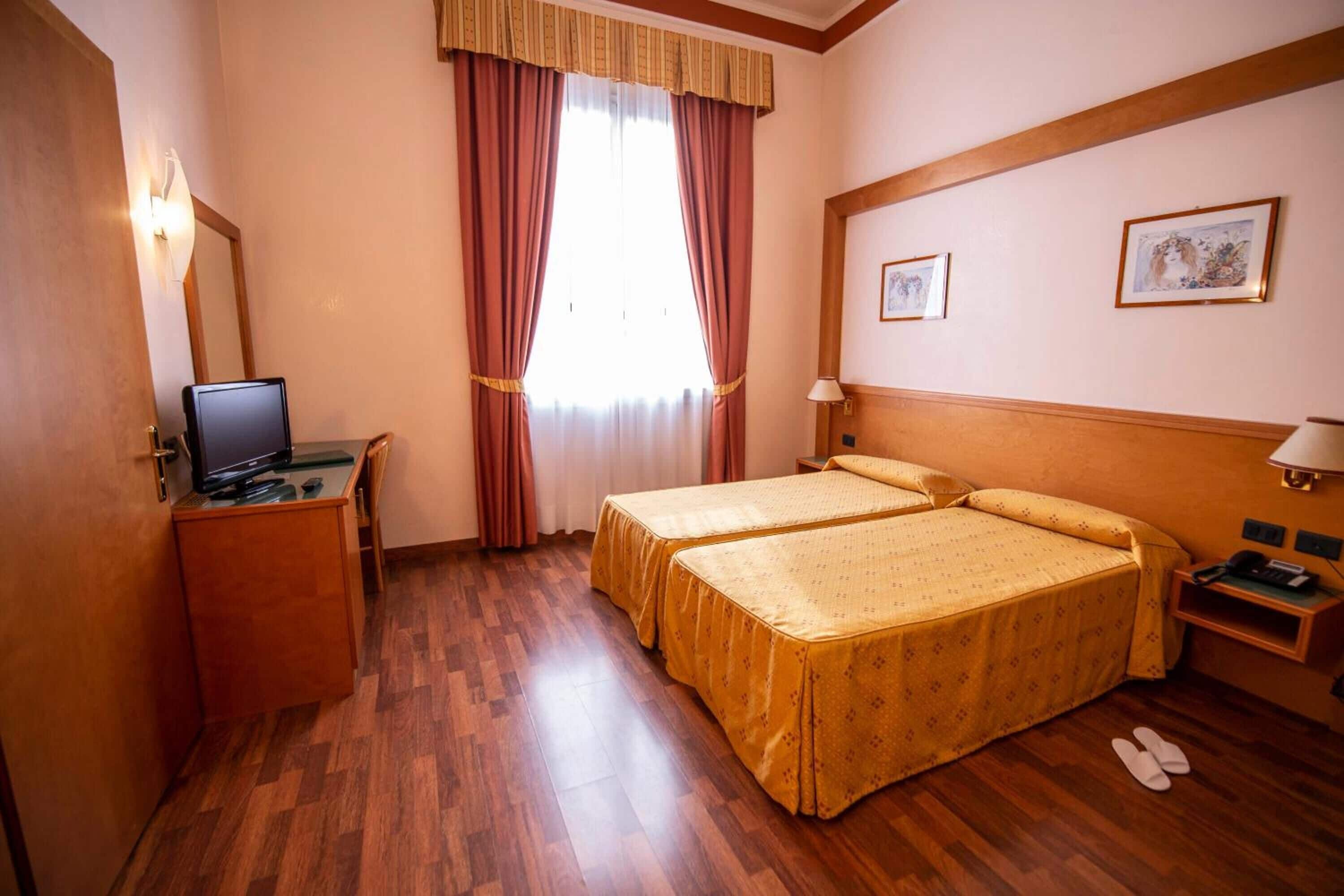 Habitación de hotel con dos camas, un escritorio con computadora, televisor, ventana con cortinas y lámpara de pared.