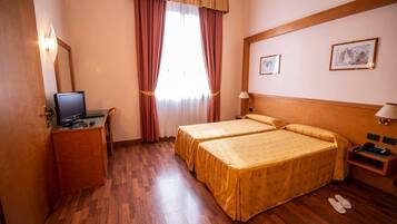 Habitación de hotel con dos camas, un escritorio con computadora, televisor, ventana con cortinas y lámpara de pared.