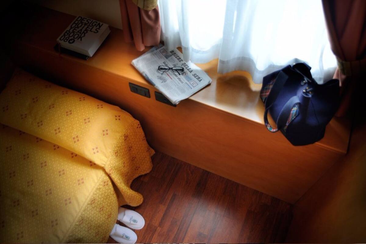 A small, cozy room with a wooden bench, a yellow blanket, a pair of slippers, a book titled 'Design Hotels Book 0800', a newspaper, and a handbag.