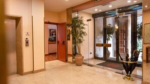 Un hall d’hôtel comprenant un comptoir d’accueil, une grande plante en pot, une porte d’entrée en verre et un ascenseur.