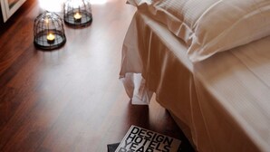 Un lit bien fait, avec une couette blanche et des oreillers, deux lampes au sol qui ressemblent à des bougies, et un magazine intitulé « LES MEILLEURS HÔTELS DE L’ANNÉE » posé sur le sol.