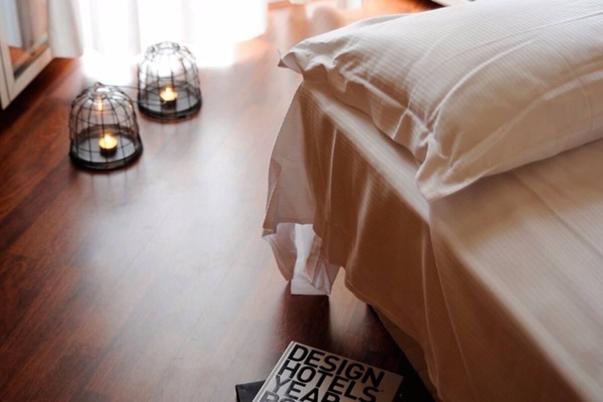 A neatly made bed with a white comforter and pillows, two candle-like light fixtures on the floor, and a magazine titled "DESIGN HOTELS YEAR'S END" on the floor.