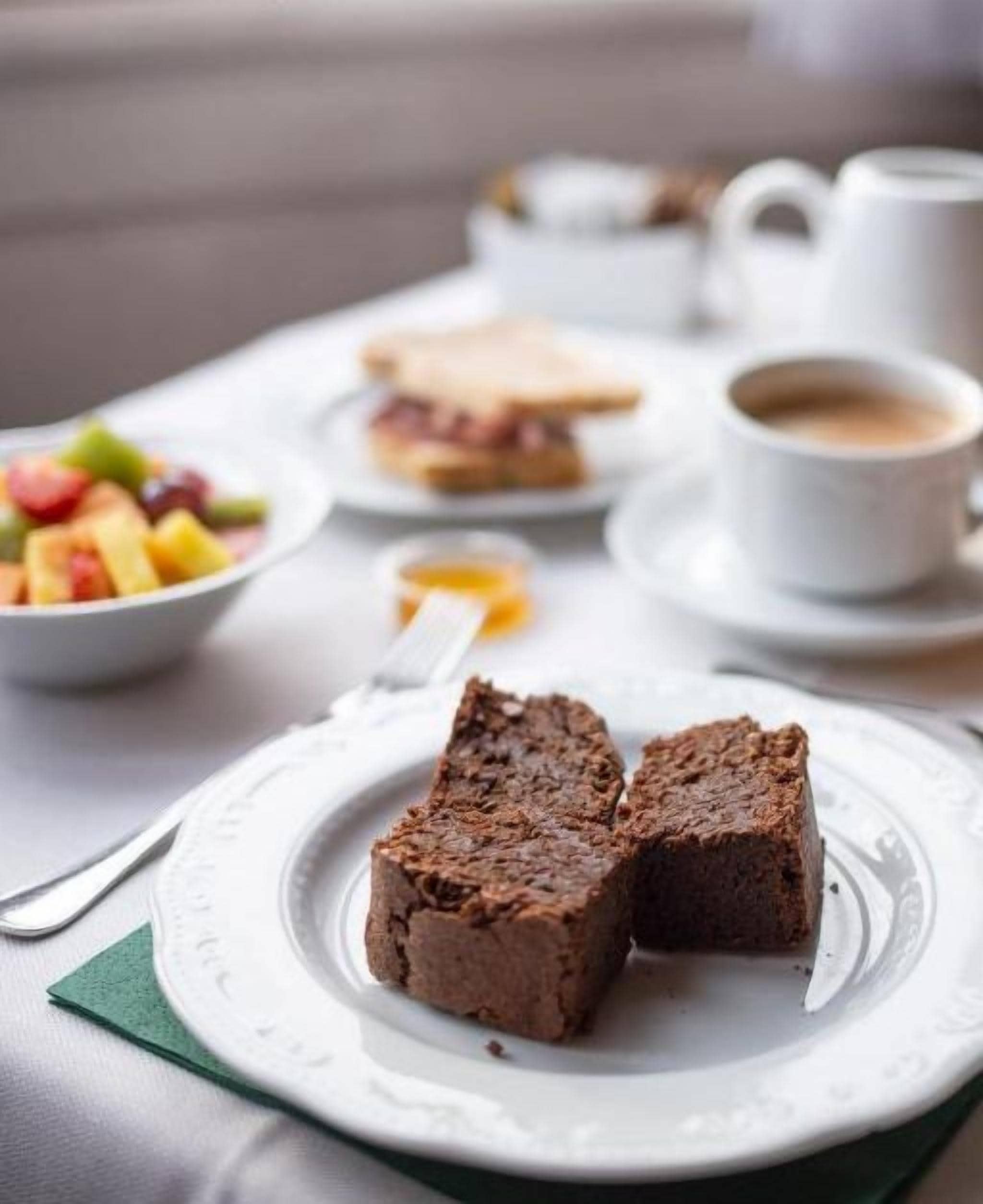Un desayuno con una fuente de brownies, un bol de fruta y una taza de café sobre una mesa.