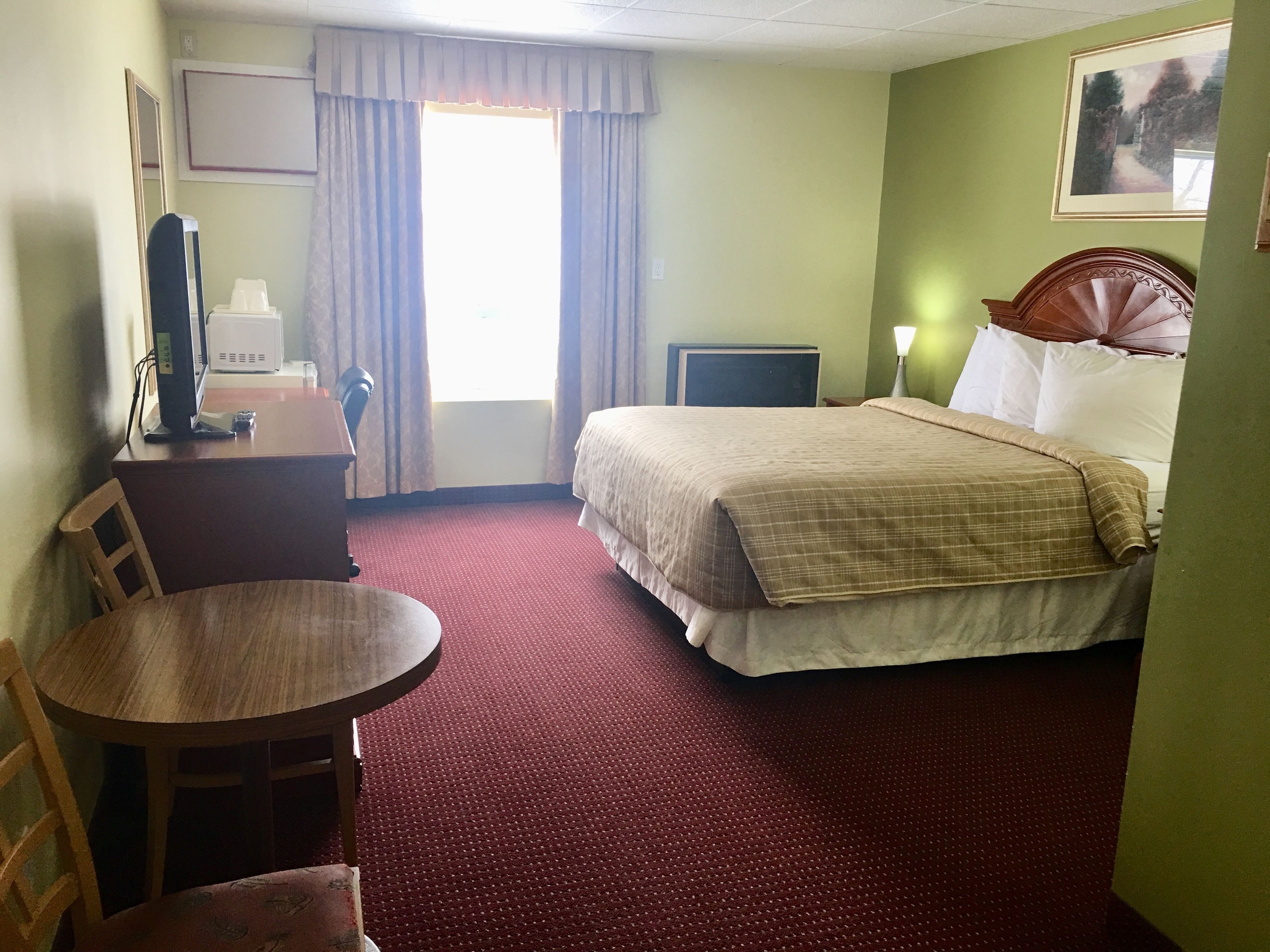 A hotel room with a bed, a desk with a computer, a chair, a small table, and a window with curtains.