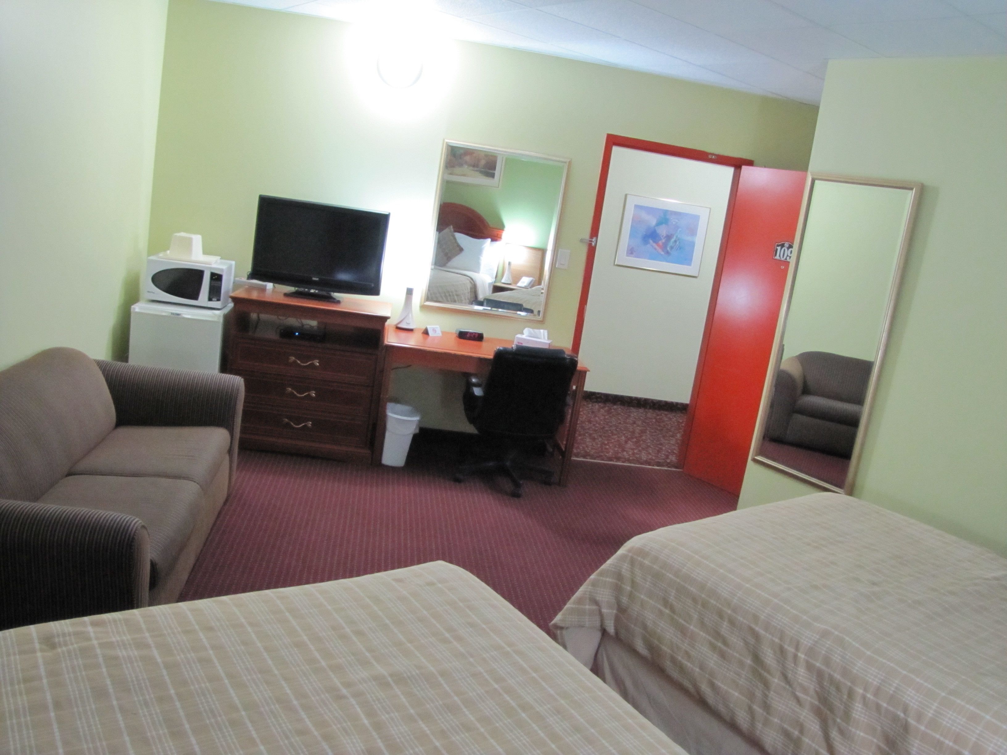 A hotel room with a bed, a desk with a computer, a chair, a microwave, and a mirror.