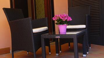 A seating area with two wicker chairs and a small table, a potted plant with pink flowers, and a door with a patterned curtain.