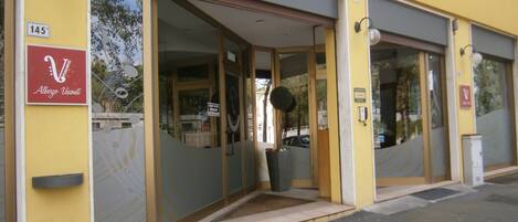 Entrada de hotel con puerta de cristal, un cartel que indica disponibilidad de habitaciones y el número 145 en la pared.