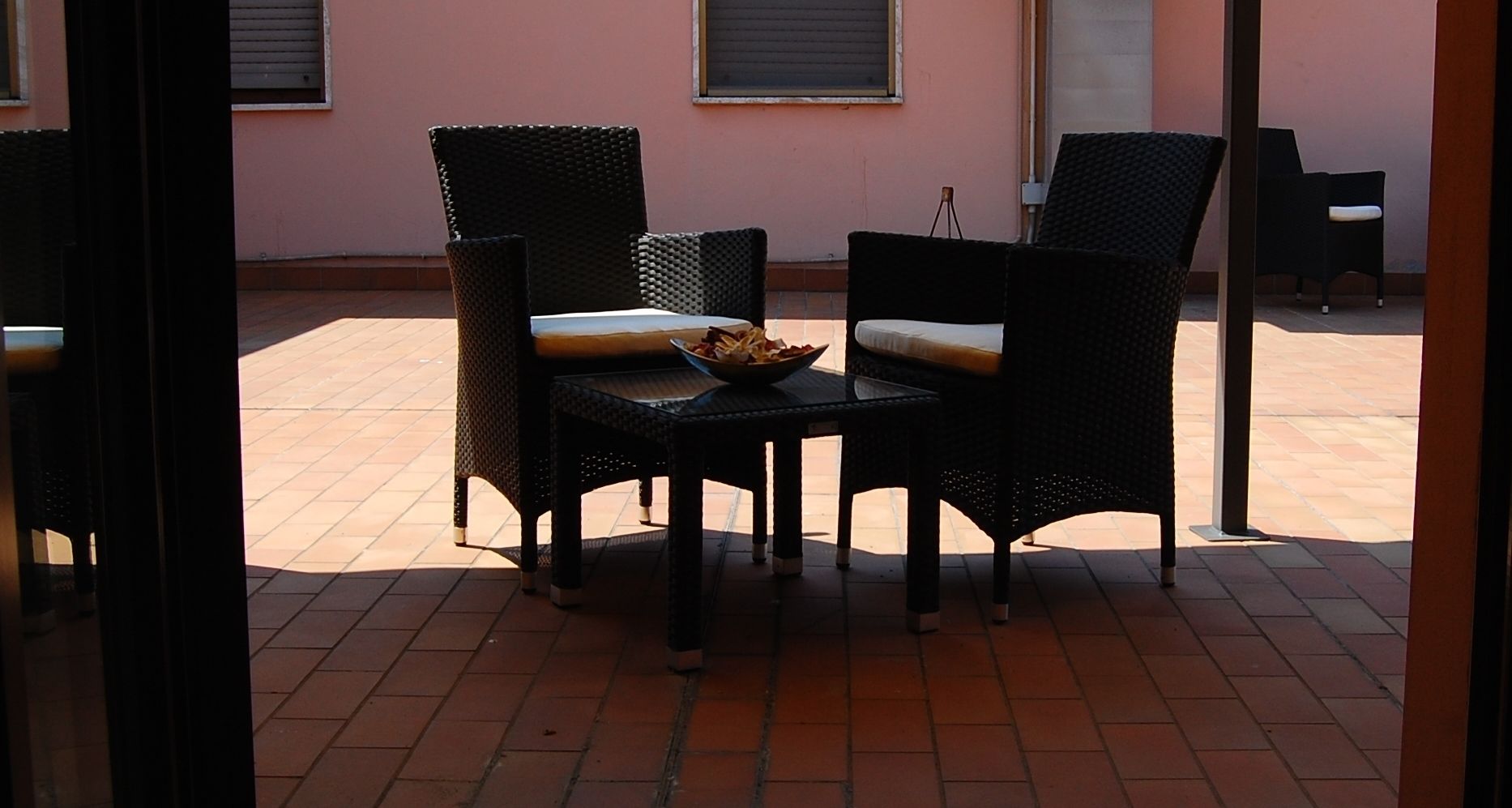 Un patio al aire libre con sillas de mimbre y una mesa, un frutero con fruta en la mesa, y un edificio rosa al fondo.