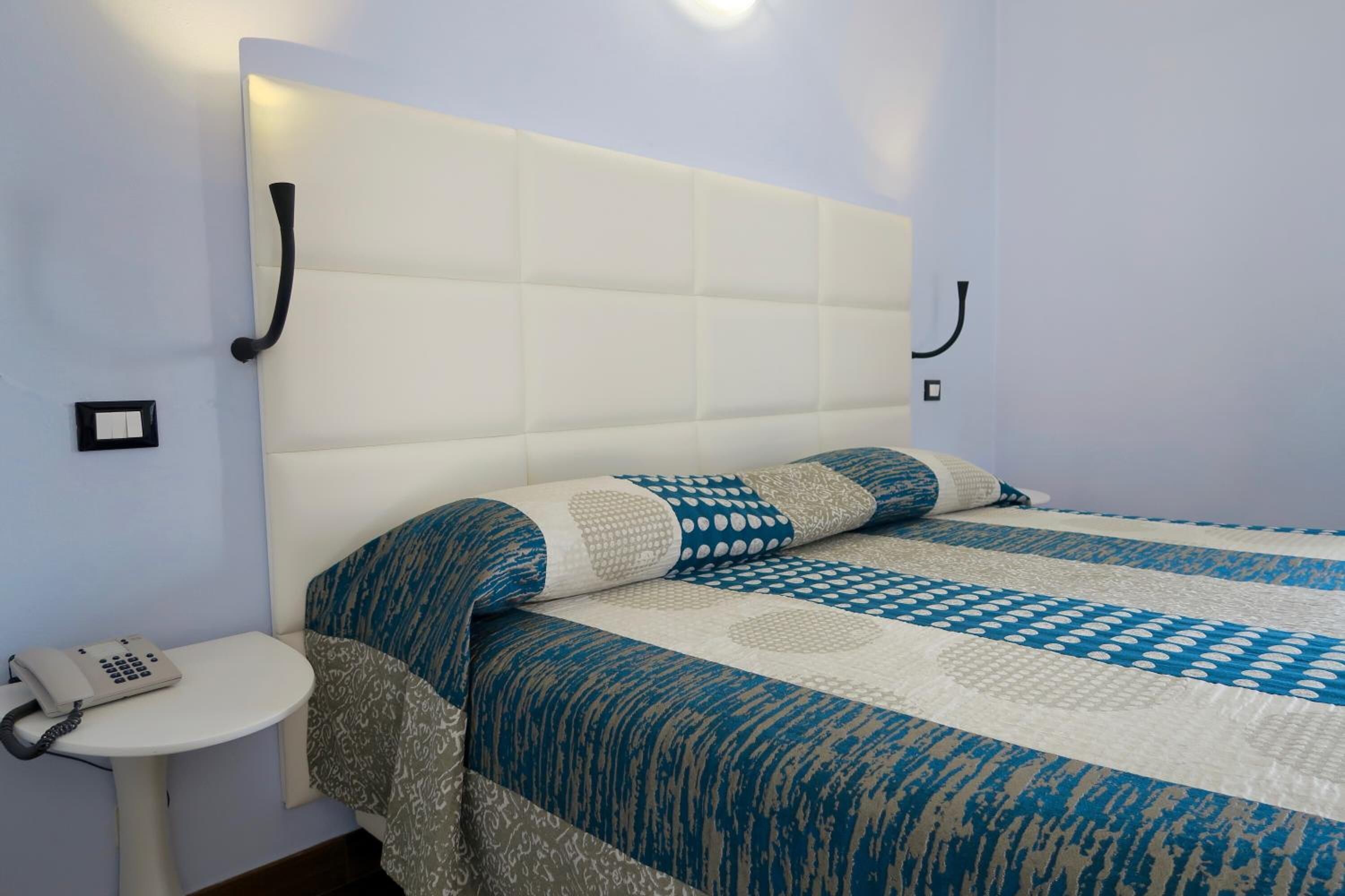 Cama bem arrumada com colcha estampada, mesa de cabeceira com telefone e luminária fixada na parede.