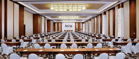 Un salón de conferencias con filas de mesas y sillas, una araña y un cartel que dice 'Atria'.