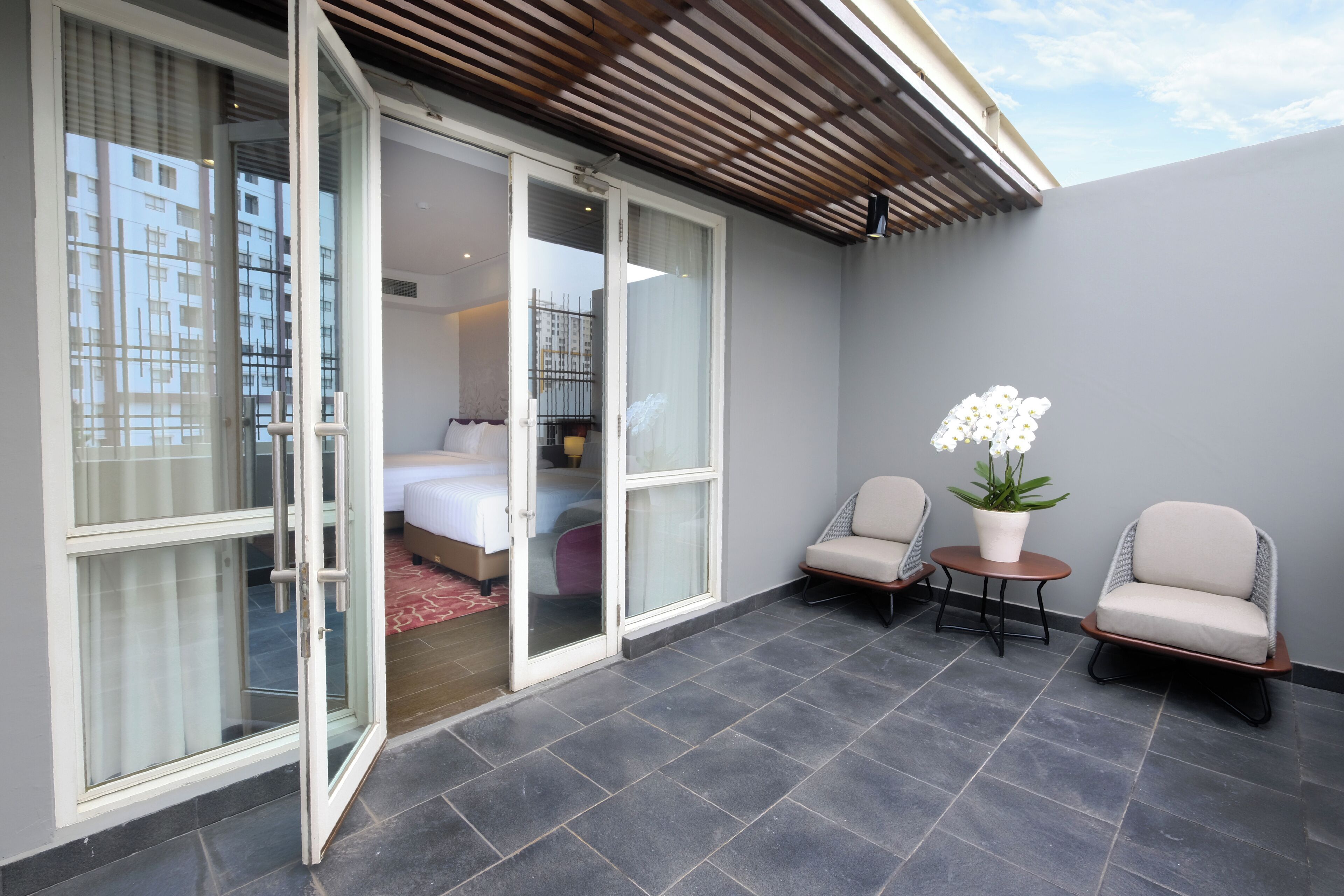 Une chambre d’hôtel moderne dotée d’une porte-fenêtre coulissante donnant sur un balcon, de deux chaises, d’une petite table et d’une plante en pot.