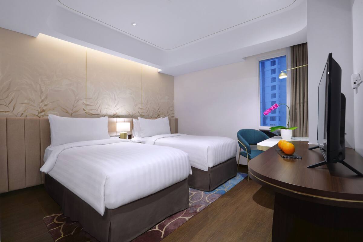 Hotelkamer met twee bedden, een bureau, een televisie en een raam met uitzicht op een gebouw.