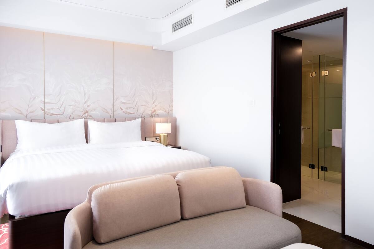 Een moderne hotelkamer met een groot bed, een bank, een nachtkastje met een lamp en een douchecabine met glazen wanden.