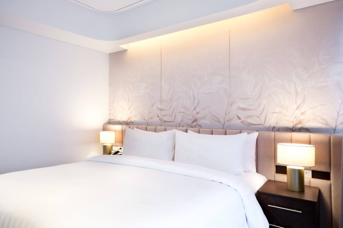 Een net opgemaakt bed met witte lakens, een hoofdbord met een bloemenprint en twee bedlampjes op een nachtkastje.