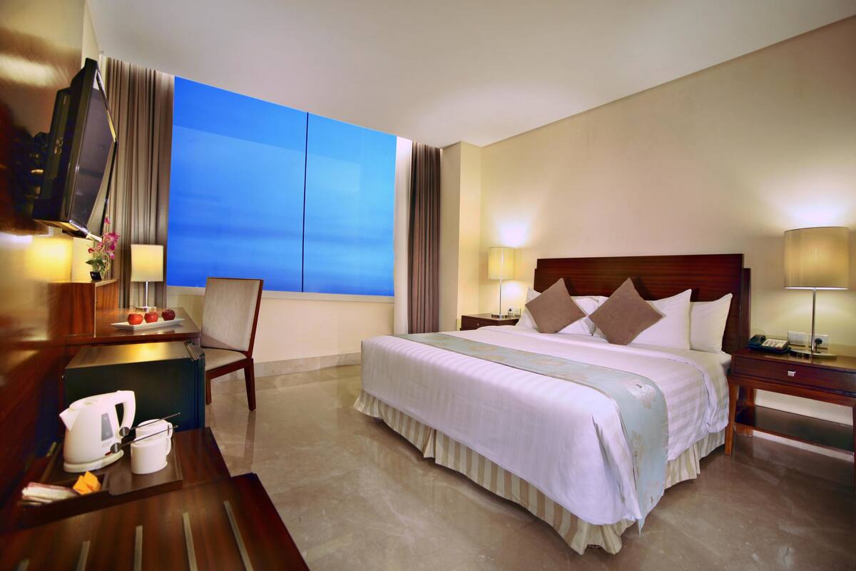 A hotel room with a large bed, a desk with a lamp, a chair, a TV, and a view of the sky.