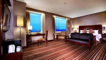 A hotel room with a large bed, two chairs, a desk with a lamp, a coffee maker, and a view of the ocean.