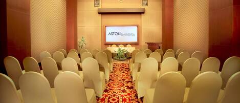 A conference room with rows of white chairs, a red carpet, and a screen displaying 'ASTON SAMARINDA'.