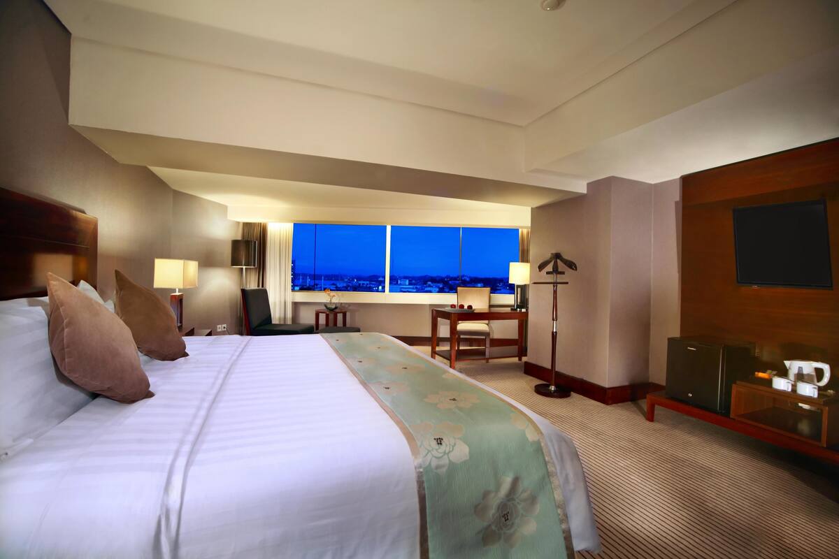A hotel room with a large bed, a desk, a chair, a television, and a view of the city at dusk.