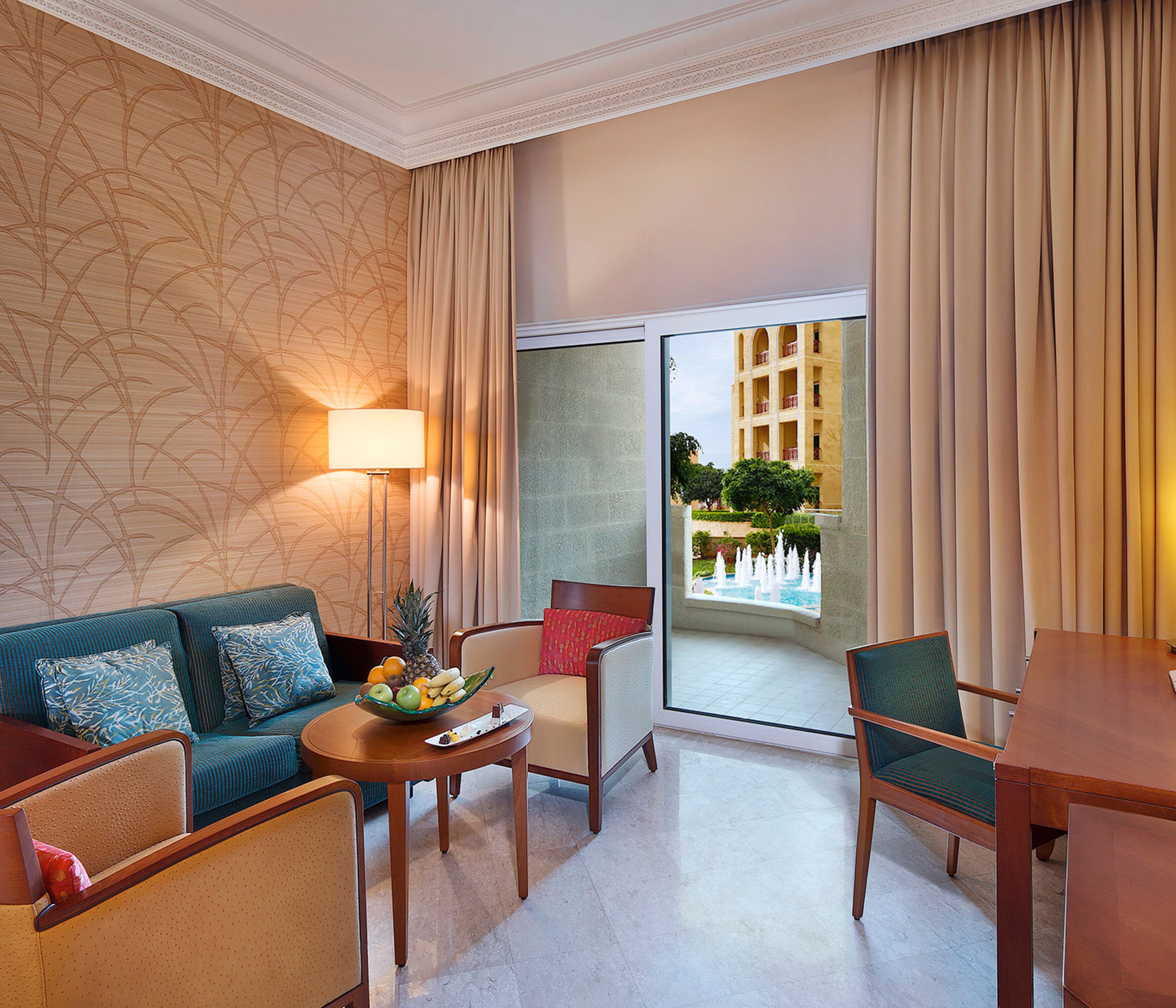 Une chambre d’hôtel avec un canapé, des fauteuils, une table avec des fruits, une lampe et une vue sur un bâtiment et un élément décoratif aquatique à l’extérieur.