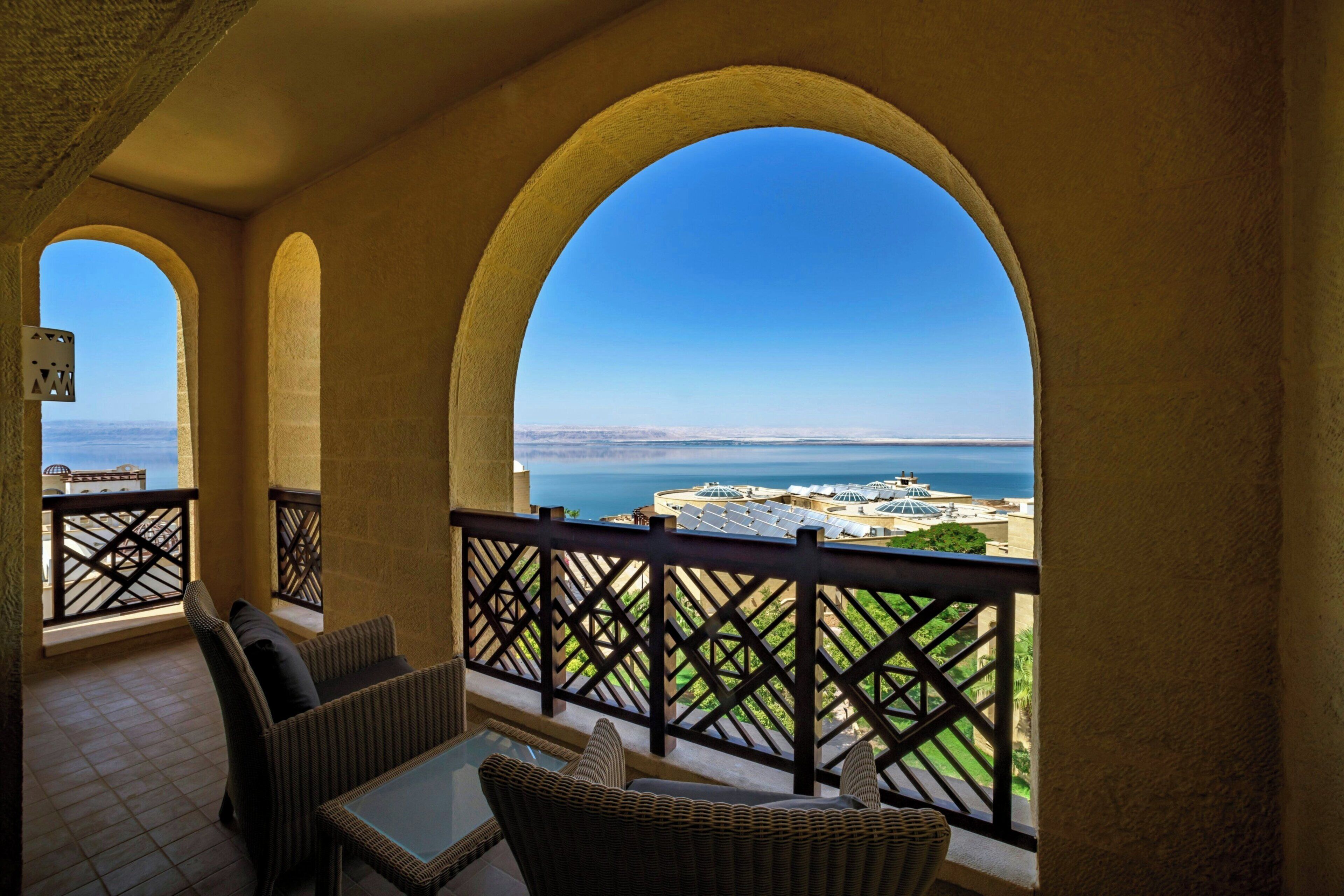 A balcony with wicker furniture, a glass table, and a view of the sea and distant buildings.
