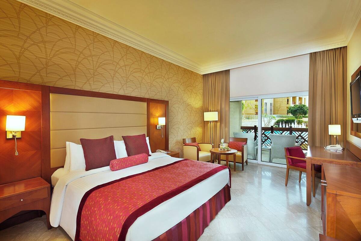 Habitación de hotel con una cama grande, un escritorio, una silla, una mesita y un balcón con vista a la piscina.